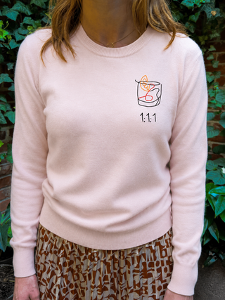 A person wearing a pale pink crewneck sweater with a small graphic of a coffee cup and the text '1.1.