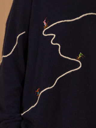 
A woman wearing a dark navy blue crewneck sweater with abstract white line embroidery and small colorful bird motifs, paired with navy pants, standing against a light wood-paneled background NAVY