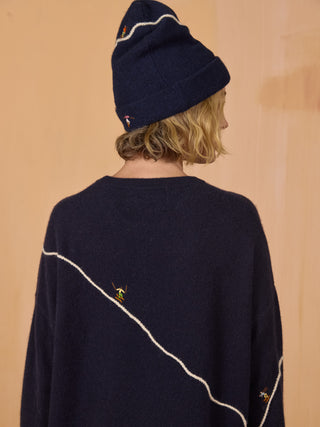 
A woman wearing a dark navy blue crewneck sweater with abstract white line embroidery and small colorful bird motifs, paired with navy pants, standing against a light wood-paneled background NAVY