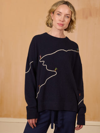 


A woman wearing a dark navy blue crewneck sweater with abstract white line embroidery and small colorful bird motifs, paired with navy pants, standing against a light wood-paneled background NAVY