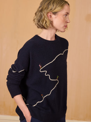 
A woman wearing a dark navy blue crewneck sweater with abstract white line embroidery and small colorful bird motifs, paired with navy pants, standing against a light wood-paneled background NAVY
